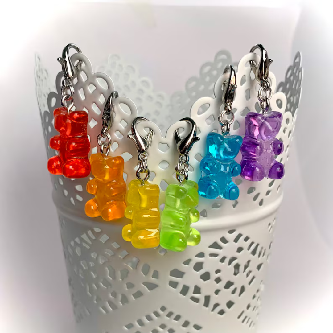 Six translucent gummy bear stitch markers hang off the side of a white cup. They are arranged in the colors of the rainbow.