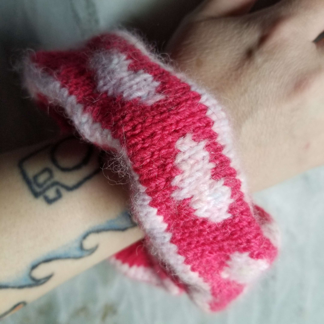 A handknit pink scrunchie worm on someone's wrist. There are hearts along the scrunchie in light pink, and the background is a dark pink.