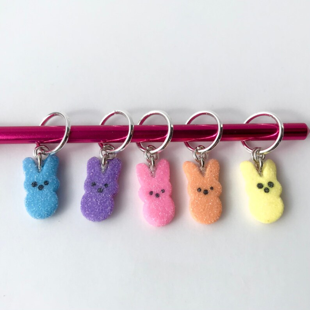 Five polymer clay marshmallow bunny stitch markers hang on a red knitting needle.
