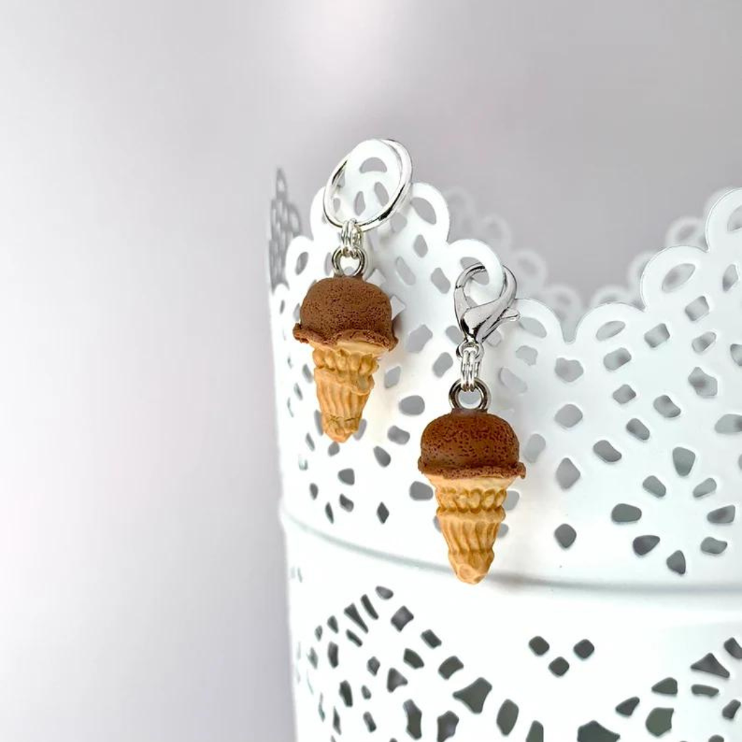 Two polymer clay chocolate ice cream cone stitch markers hang on the side of a white cup.