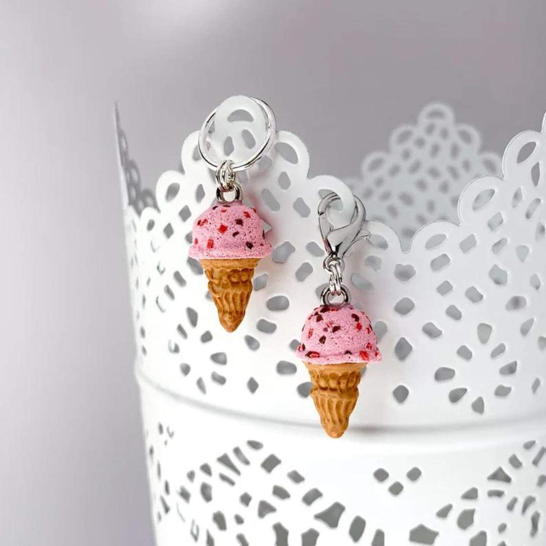 Two polymer clay strawberry ice cream cone stitch markers hang on the side of a white cup.