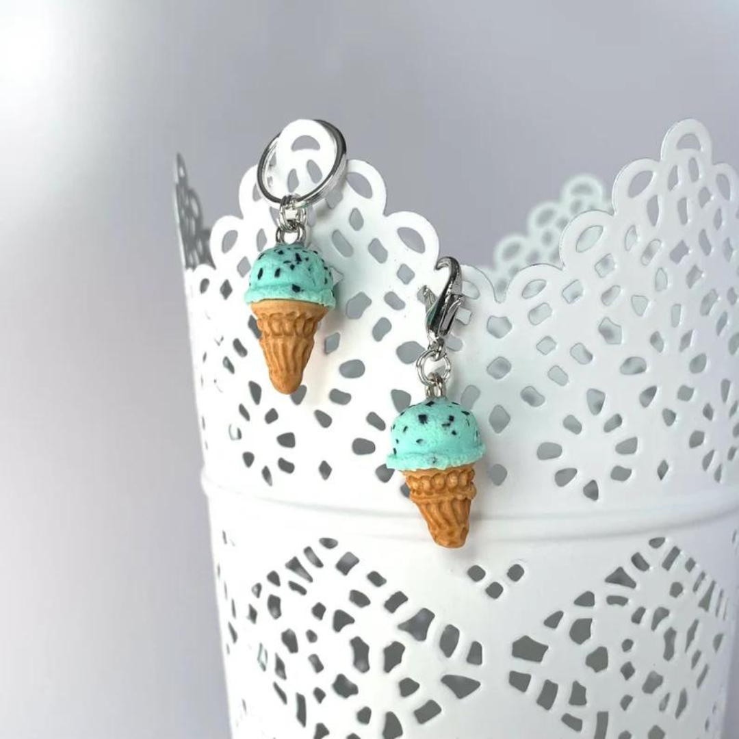 Two polymer clay mint chocolate chip ice cream cone stitch markers hang on the side of a white cup.