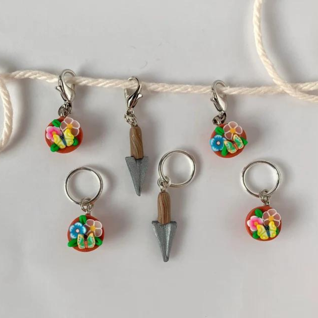 Polymer clay stitch markers arranged flat on a table with string. Four are small pots with various flowers. Two are small garden trowels.