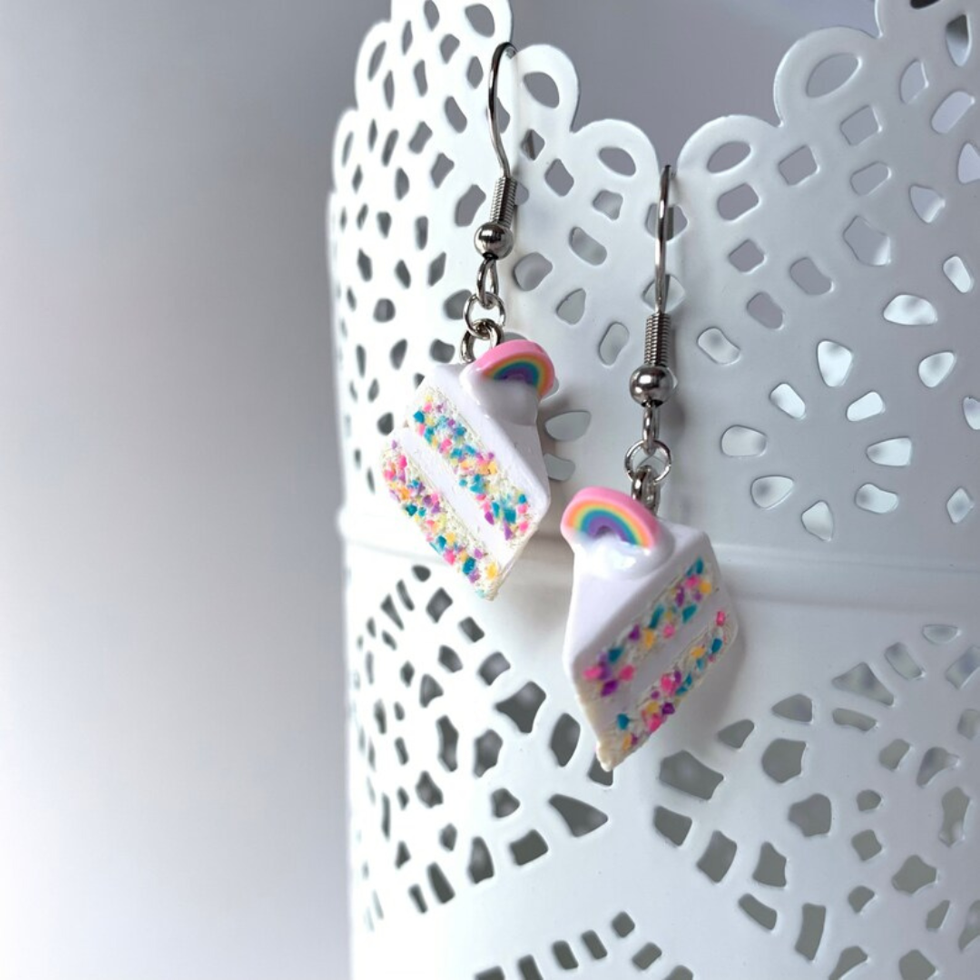 Two polymer clay vanilla birthday cake earrings hang on the side of a cup.