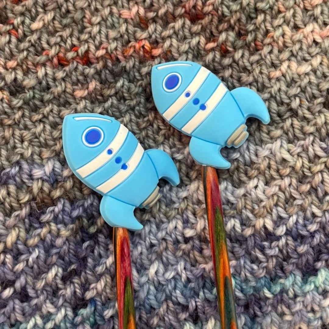 Blue rocketship knitting needle stoppers on the ends of knitting needles.