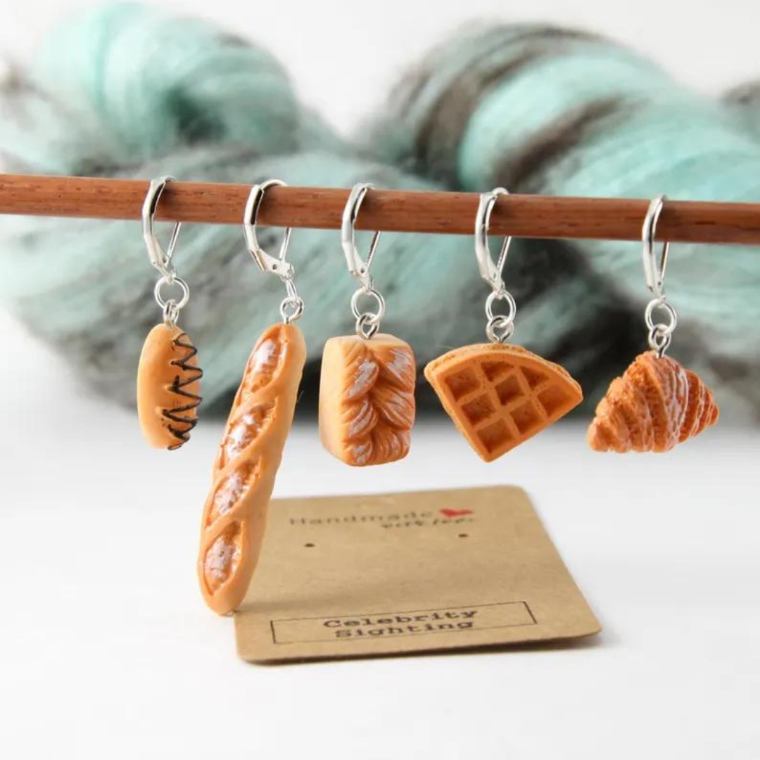 Five small bread themed stitchmarkers hanging on a wooden rod.