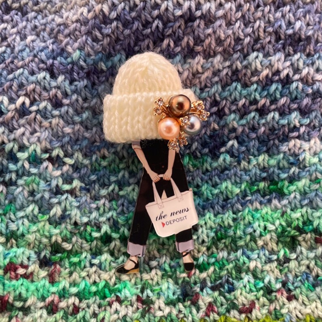 Handmade knitter's brooch with pearl accent and small handknit hat, pinned on a handknit variegated fabric.