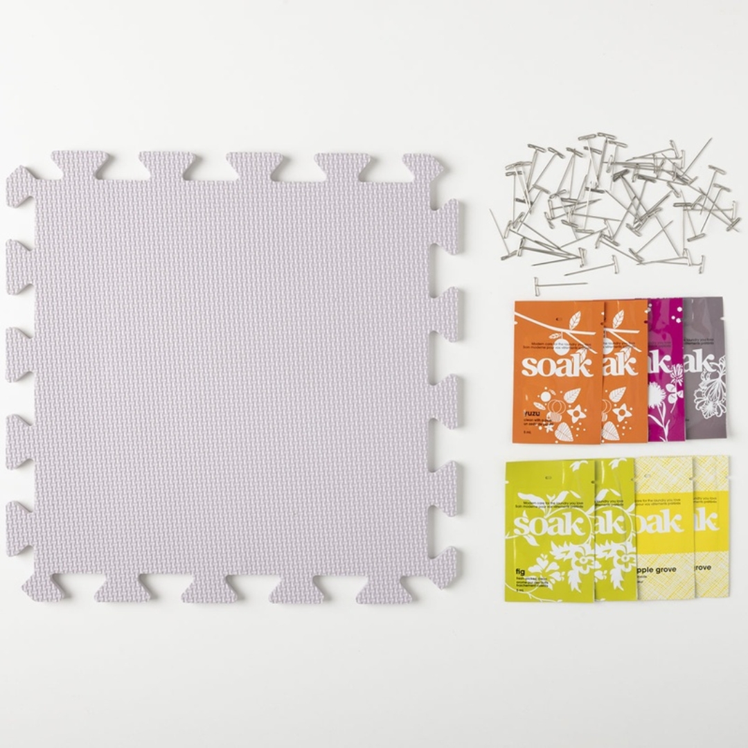 A flatlay of a blocking set. It shows a blocking mat next to a set of T-pins, and eight packets of soak wash.