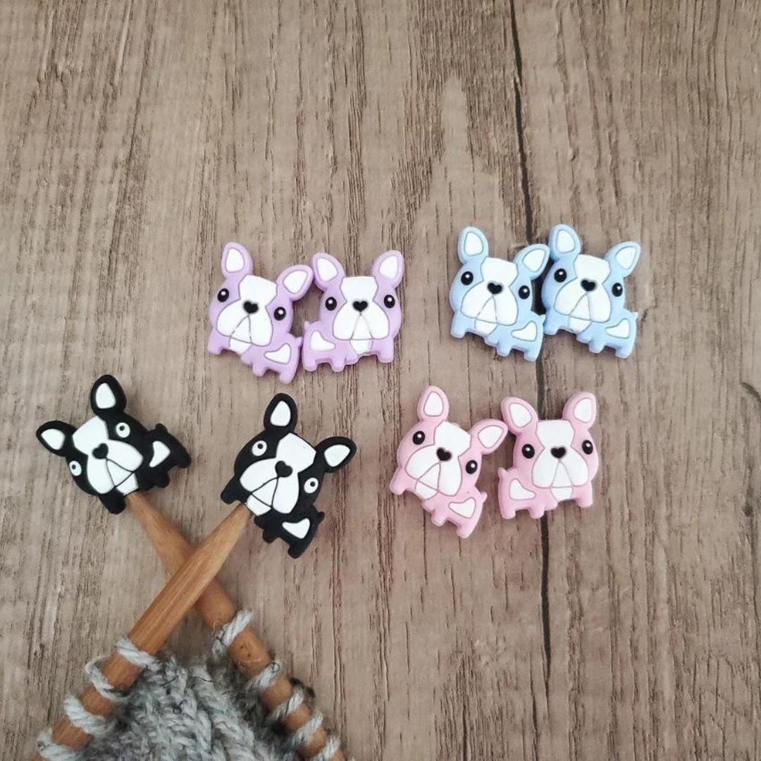 A set of black bulldog point protectors on a pair of knitting needles with a project on them. On the table next to them there are three sets of the same point protectors in purple, blue, and pink.