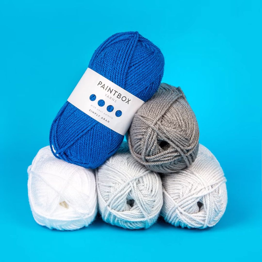 Five skeins of yarn in shades of grey and blue. Three lay on the bottom layer, with one on top turned to show a Paintbox Simply Aran label.