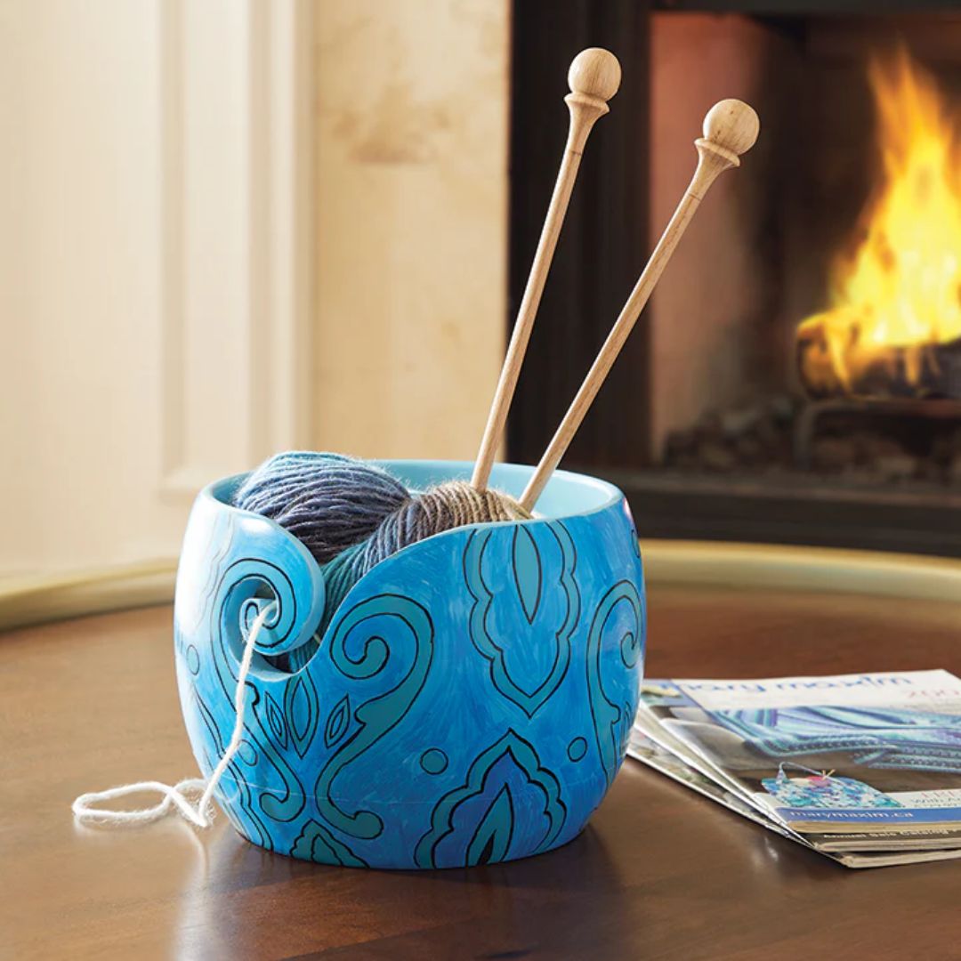 A bright blue yarn bowl with green leafy designs pictured on a coffee table in front of a fireplace.