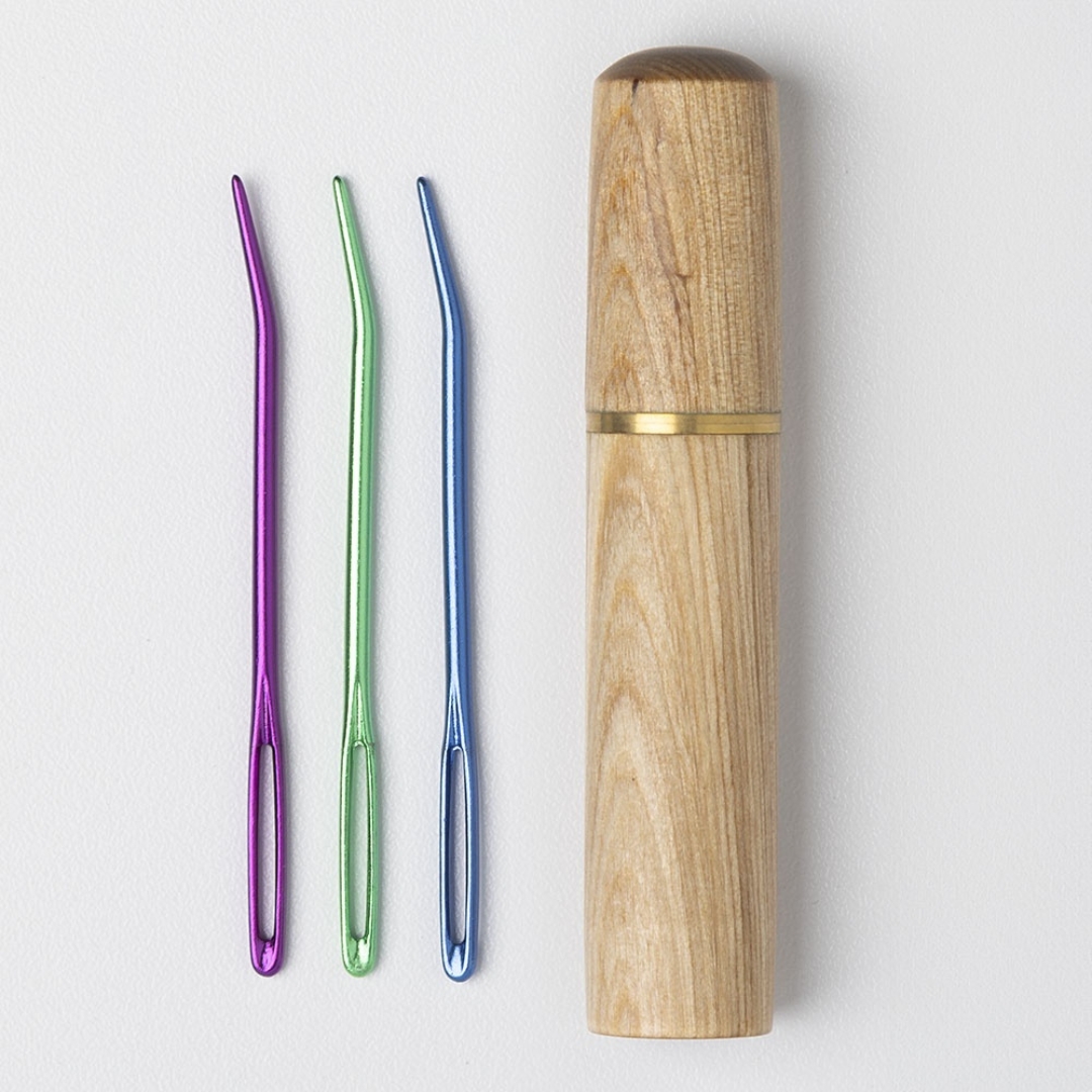A set of three bent-tip tapestry needles in purple, green, and blue. Next to them is a wooden needle holder.