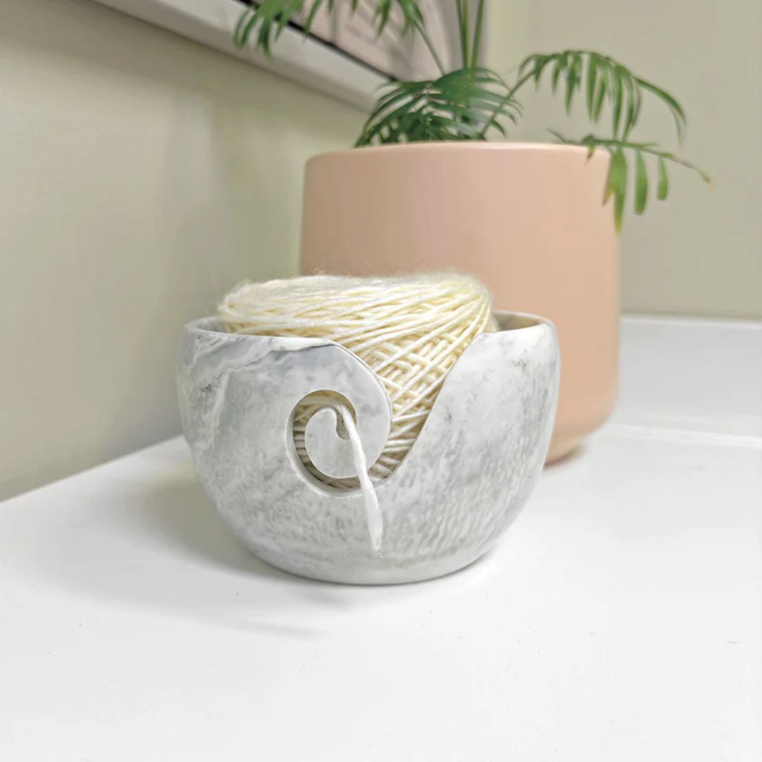 A grey and white marble resin yarn bowl, pictured with a plant behind it.