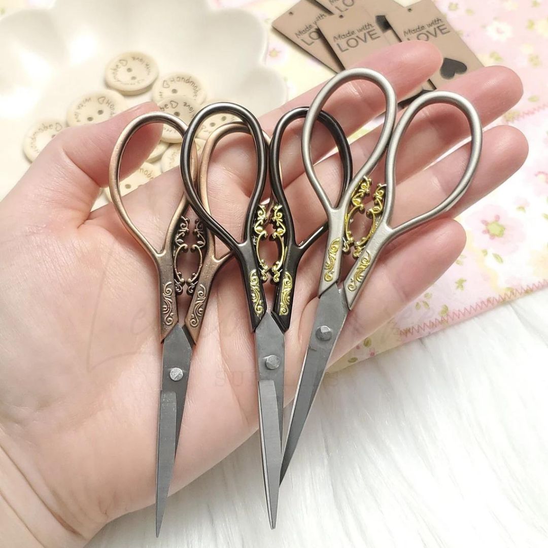 A set of three yarn scissors in someone's hand. They each have small metal embellishments on the handle. From left to right, the handles of each are rose gold, black, and silver.