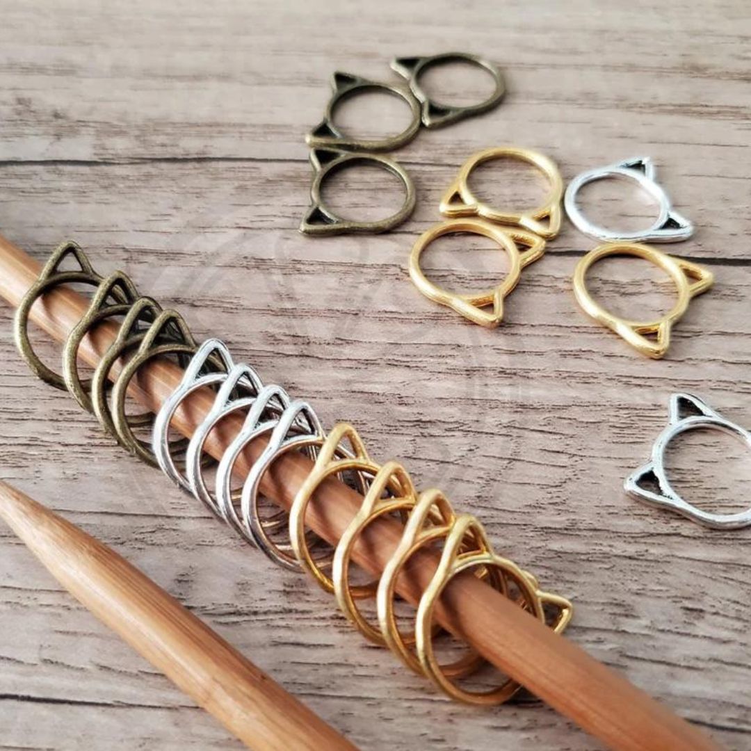 A knitting needle with black, silver, and gold cat head metal stitch markers on it. More of the same stitch markers are scattered on the table next to it.
