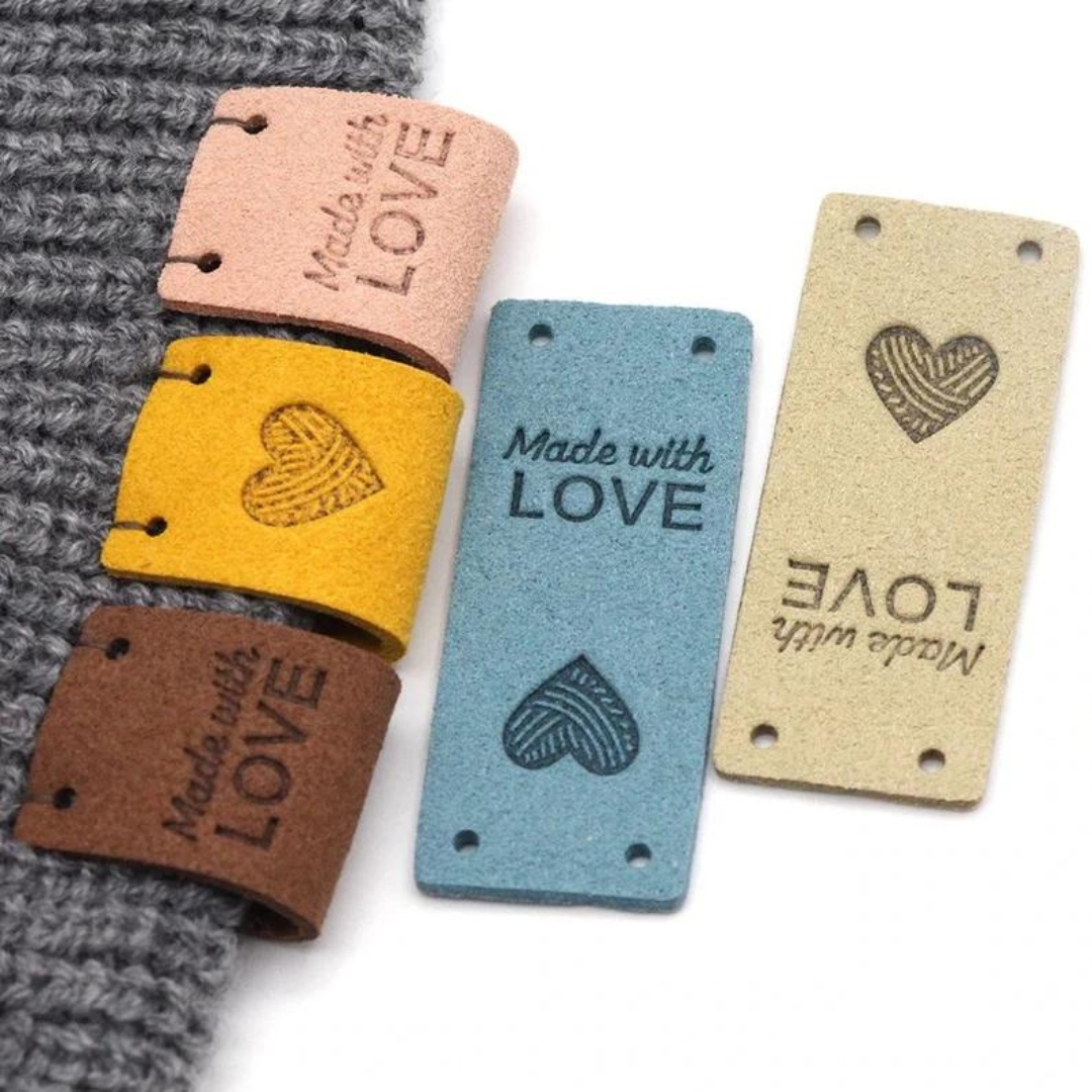 A set of rectangular leather labels. Three are sewn onto a knit swatch, two are laid flat. They all say "handmade with love" and have a heart on the bottom.